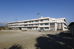 中央市立田富中学校の外観写真。グラウンドの向こうに白い校舎があり、校舎の前にはグラウンドに向かって夜間用のライトがあります。