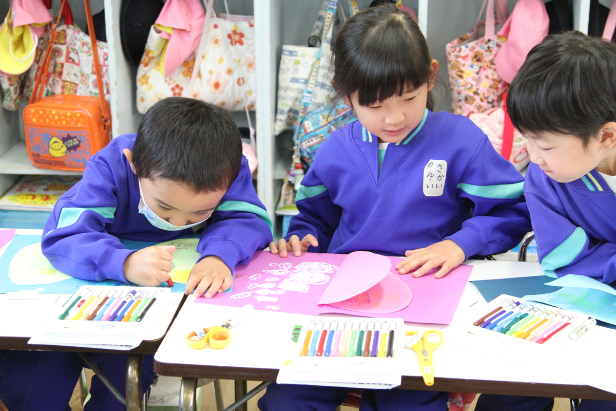 紙を手でおさえている女の子の園児の作品を見ている男の子と園児と絵を描いている園児が映っている様子