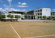 中央市立三村小学校外観。手前に大きくグラウンドが写っており、写真奥にグレーの校舎が建ち並んでいます。
