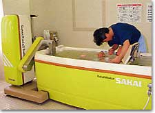 中央市立豊富デイサービスセンター内にて、入浴機能訓練をおこなっている写真。特別な機械が備えられた浴槽に高齢者の方が入浴しており、デイサービスの職員が介助しています。