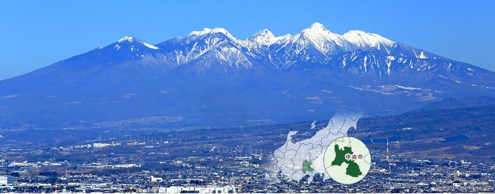 青空と雪を冠する八ヶ岳連峰のコントラストが美しい背景に中央市を眼下に望む風景。右下に山梨県の国中地方、甲府盆地の南部にある中央市の位置を指示した図。
