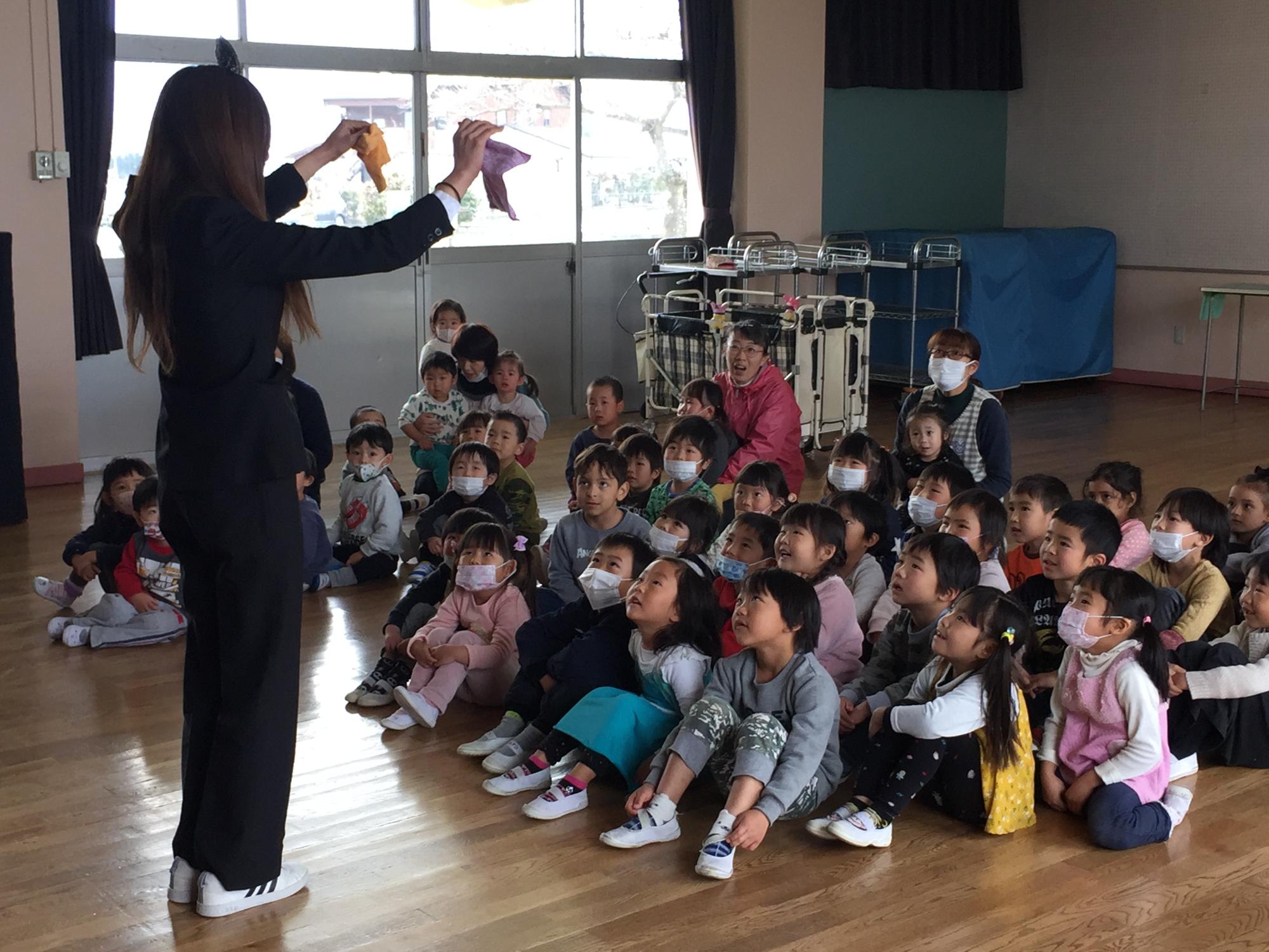 誕生会に手品を披露する保育士を見上げるように座る子どもたち
