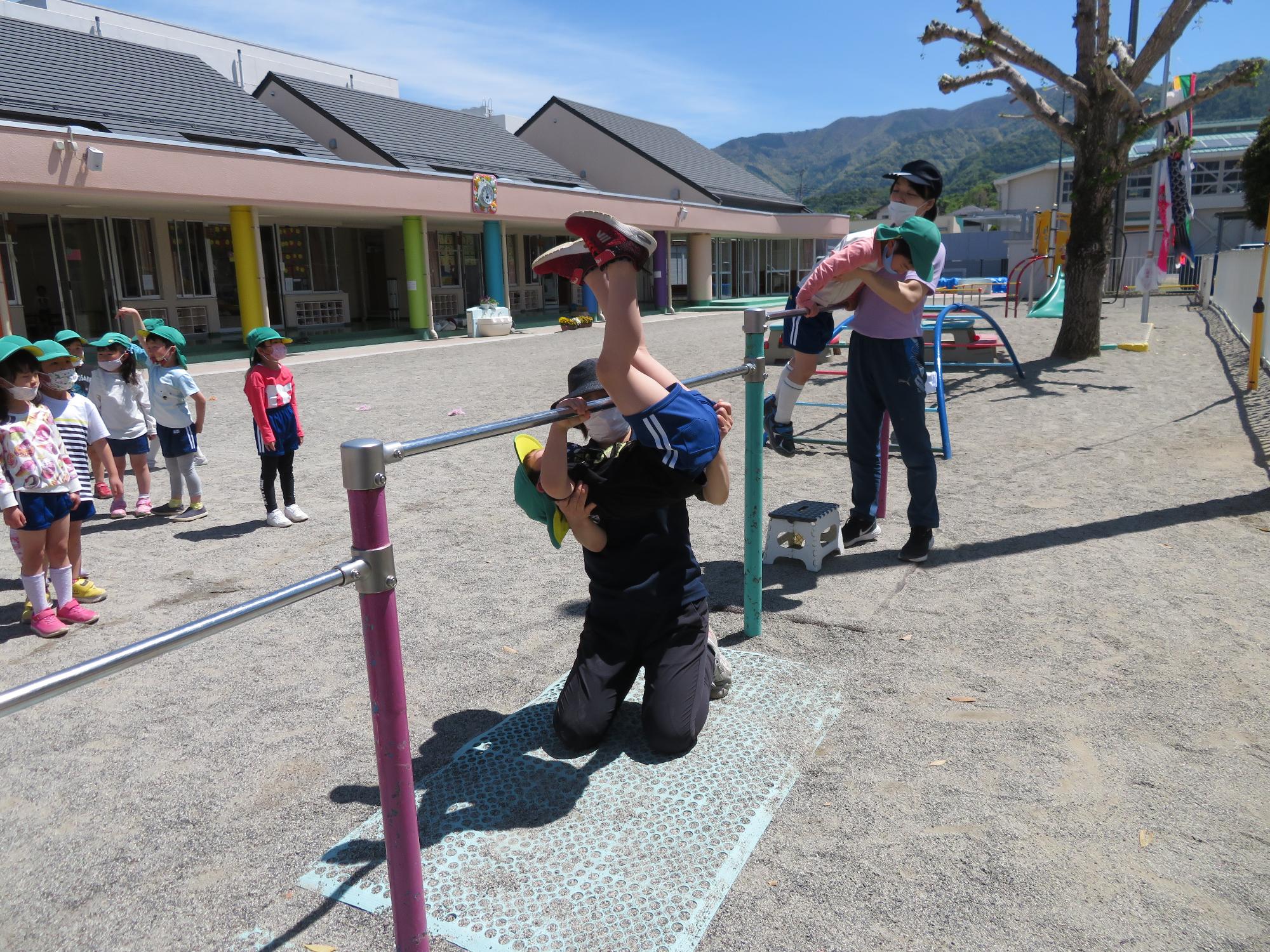 戸外で鉄棒の逆上がりを補助付きで練習する5歳児の子どもたち
